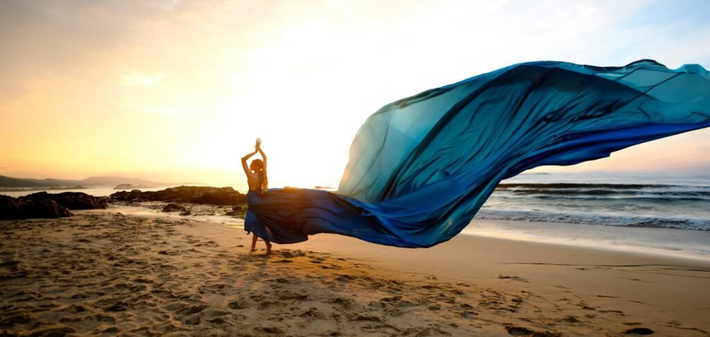 A graceful woman in a long blue dress dances on the beach during a stunning sunset.